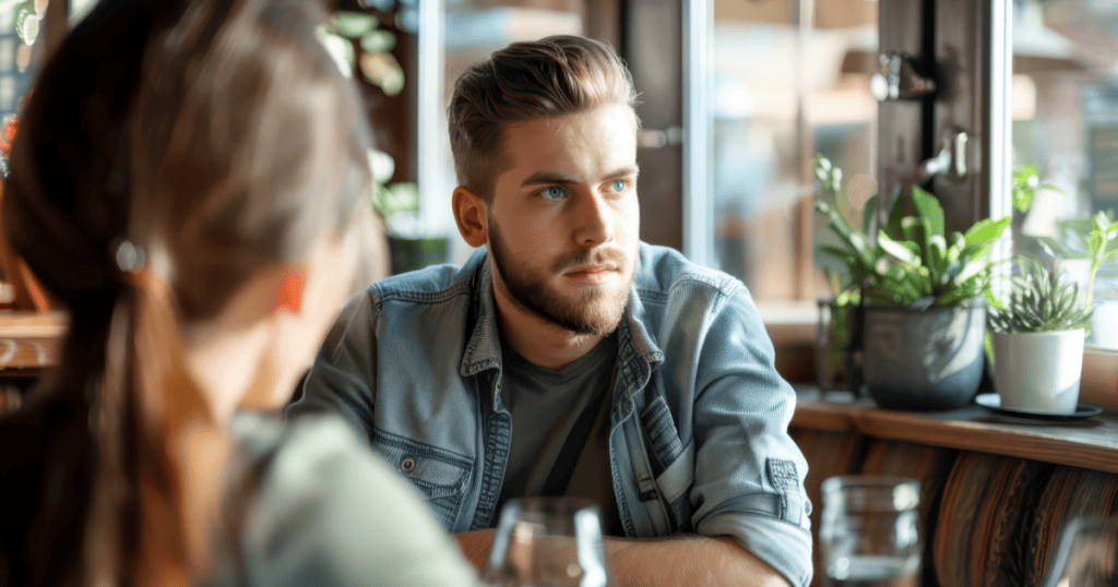 A man sits across from his partner at a café with a guarded, noncommittal stare, reflecting how a narcissist avoids introducing you to their inner circle so no one can hold them to a future with you.