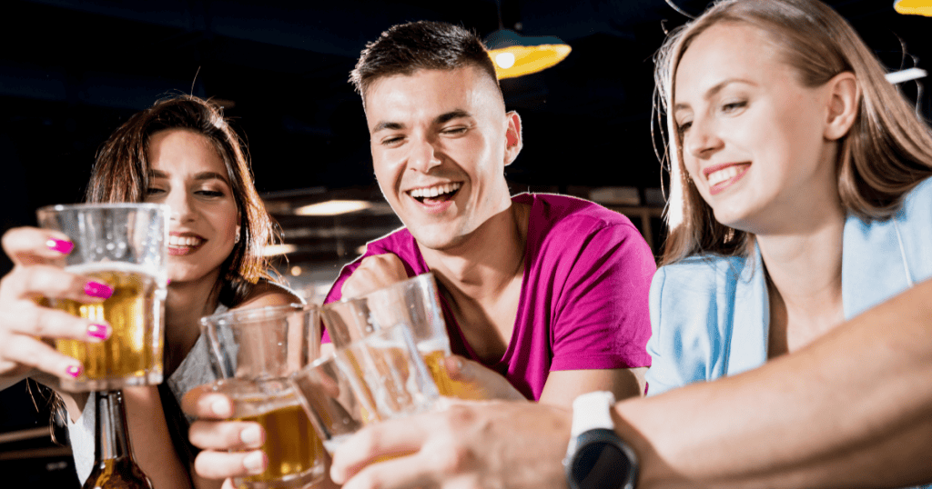A smiling man clinks glasses with two women at a lively bar, illustrating how a narcissistic partner keeps you away from their social circle so they can appear available and keep their options open.