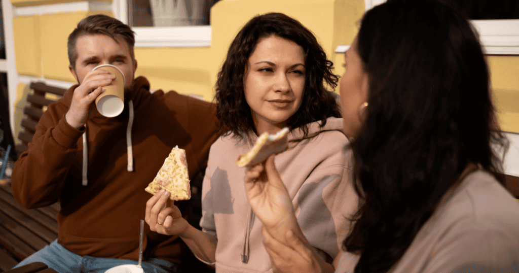 A woman eating pizza with friends gives a cool, skeptical look across the table, reflecting how a narcissist keeps forgiveness just out of reach no matter how many times you apologize.