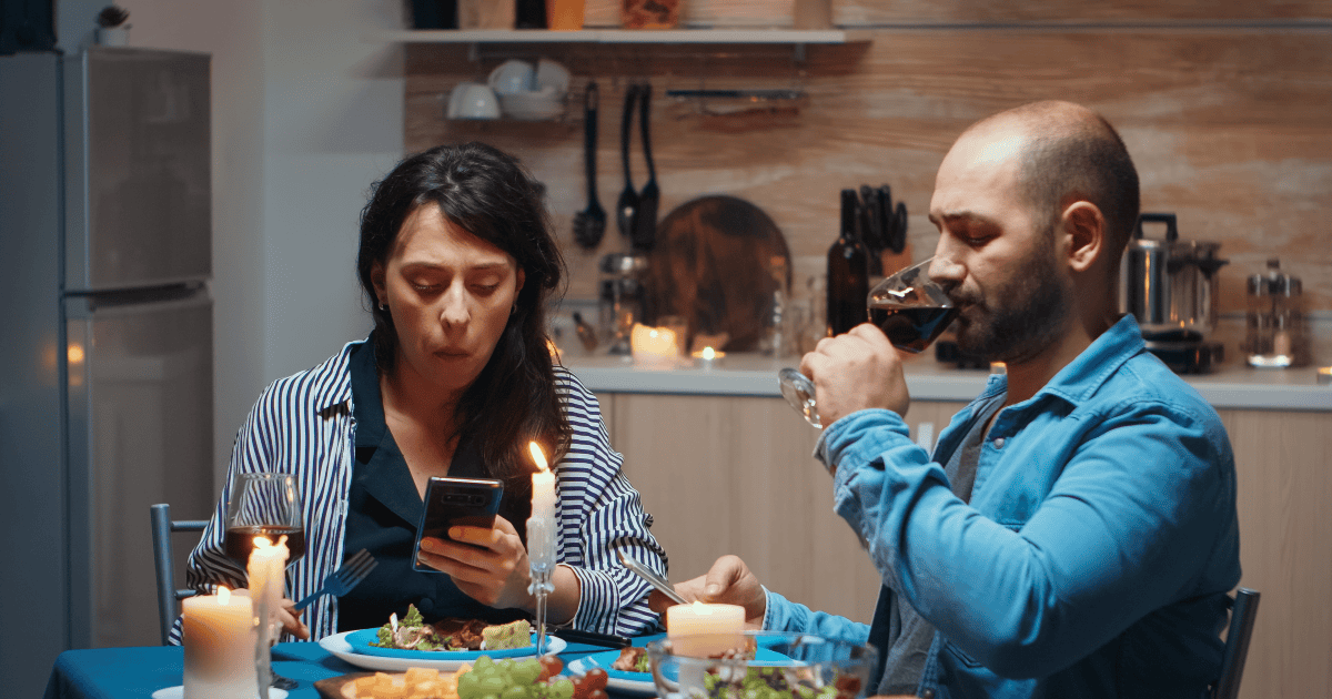 A woman reads her phone with a troubled expression during dinner, reflecting how a narcissistic parent’s hurtful messages could leave you hurting while they quietly felt they’d won.